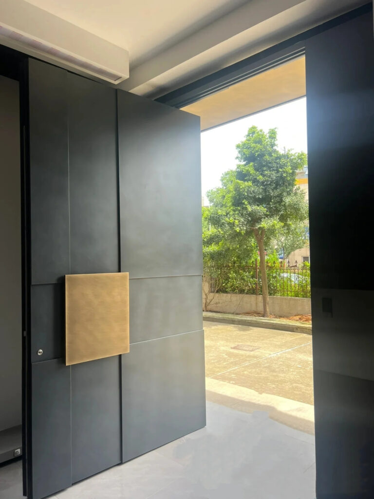 Large modern black pivot door featuring a unique oversized square gold handle plate, opening to a residential exterior.